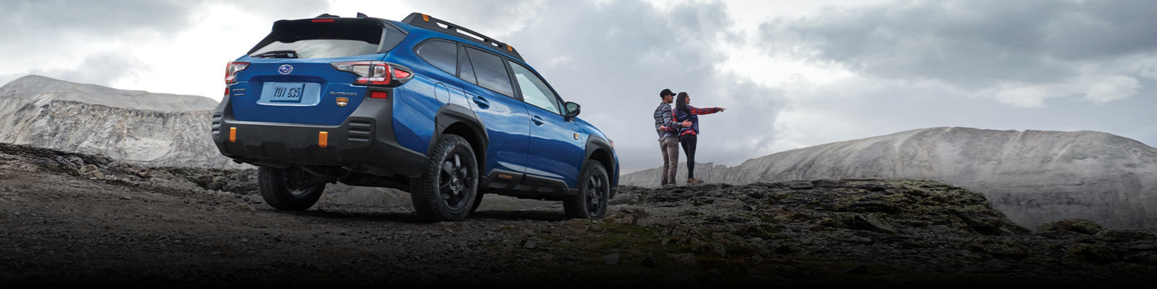 2022 Outback Wilderness in Geyser Blue parked on a rocky ridge behind a couple observing the scenery.