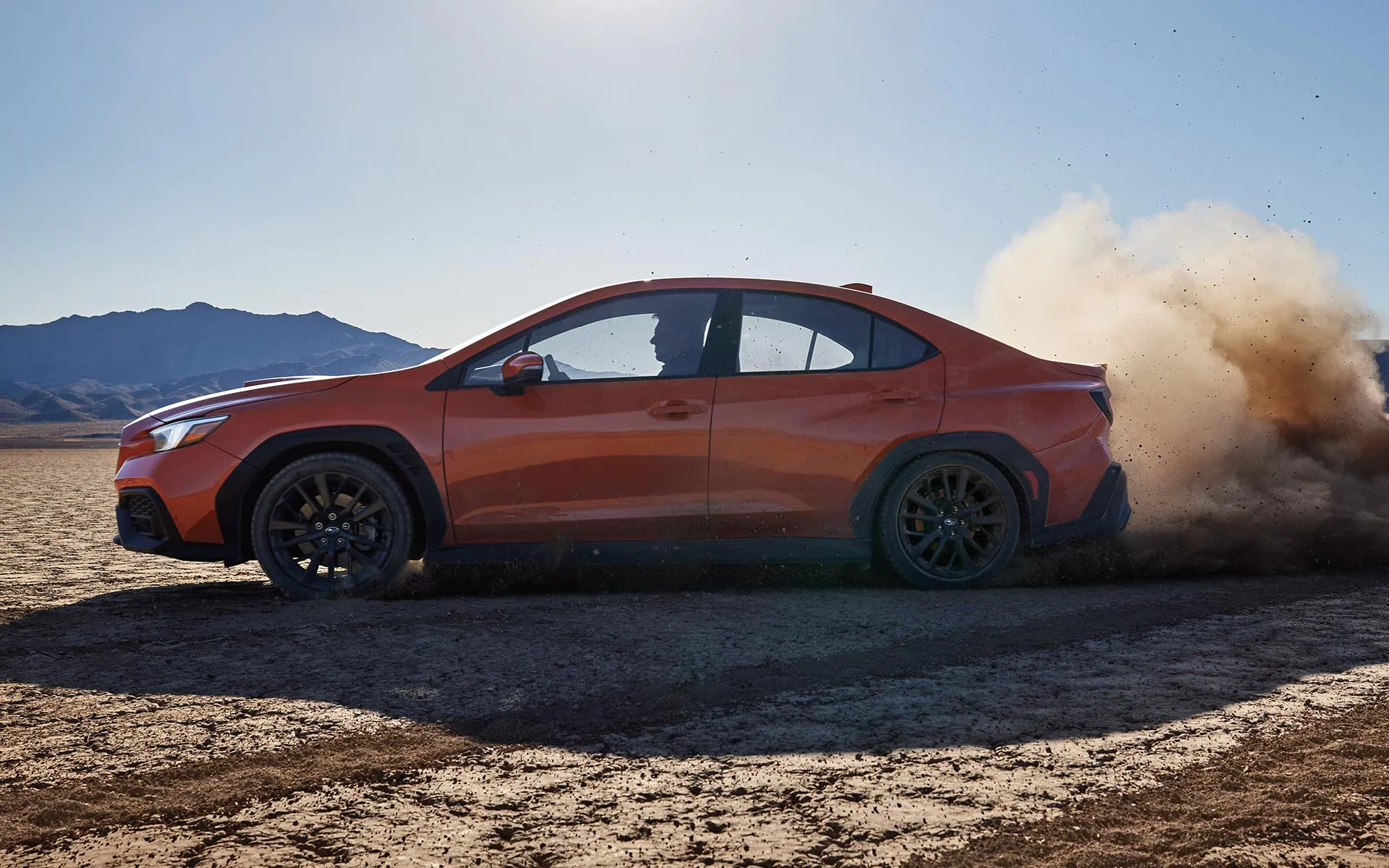 Side view of a man kicking up dust in an orange Subaru WRX